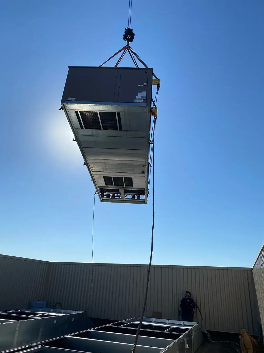 Crane lifting a large rooftop HVAC unit onto a building against a clear blue sky