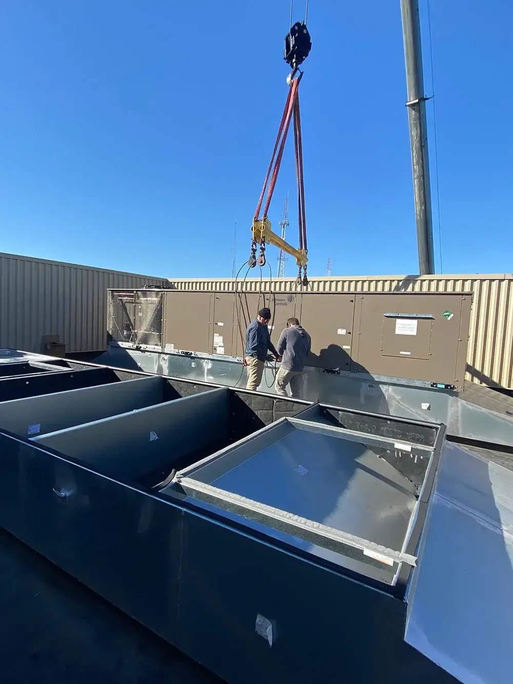 Technicians installing a large rooftop HVAC unit with a crane under a clear blue sky