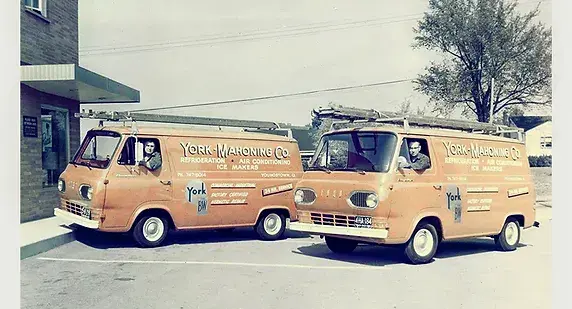 Two orange York Mahoning Co. vans parked outside a brick building. The vans have company logos and text promoting refrigeration, air conditioning, and heating services